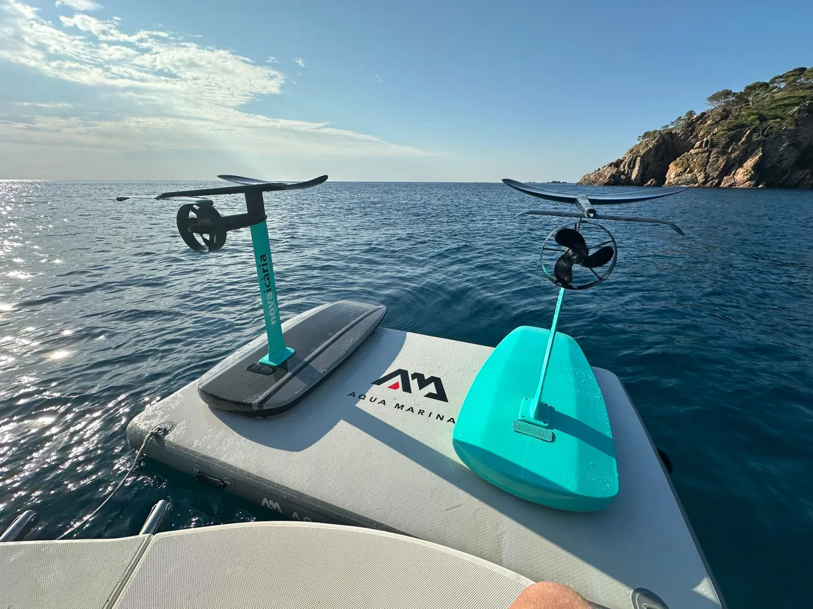 Dos tablas de e-foil listas en la plataforma del barco frente a una cala de Llafranc, Costa Brava